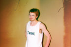 Man wearing a white tank top with 'TWINK' text against a textured wall.
