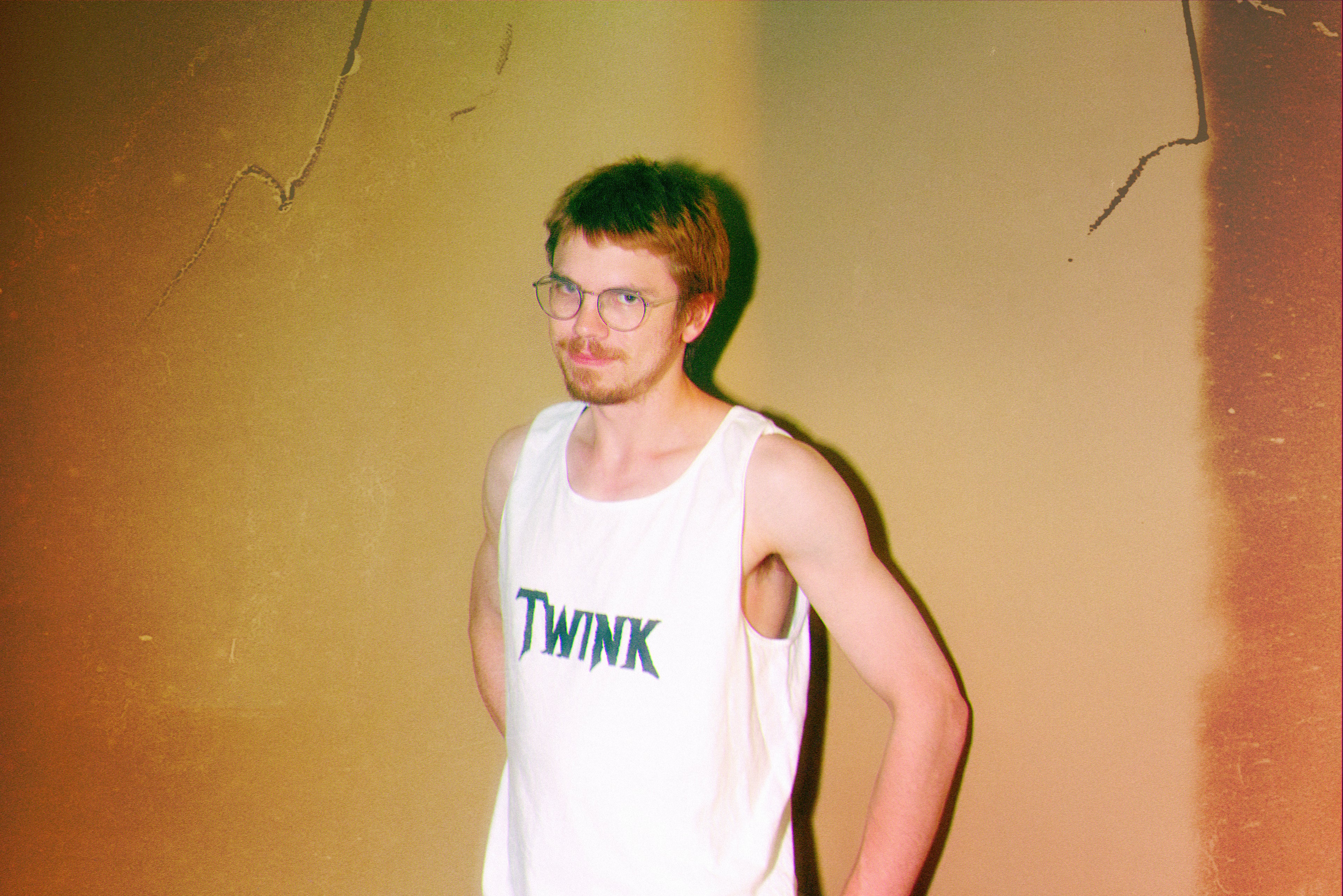 Man wearing a white tank top with 'TWINK' text against a textured wall.